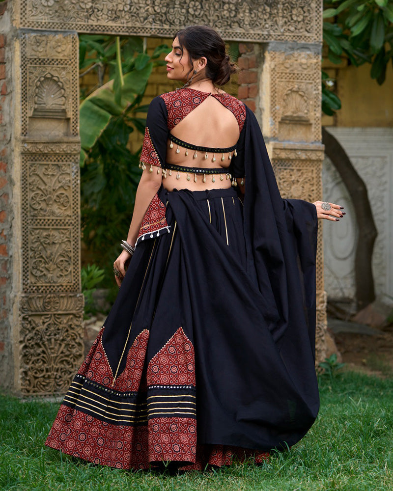 Black cotton printed lehenga choli set with traditional red and black geometric patterns, gota patti and mirror work, paired with a matching dupatta featuring decorative tassels. Ideal for festive occasions or ethnic wear collection, showcased outdoors with ornate stone carvings in the background.