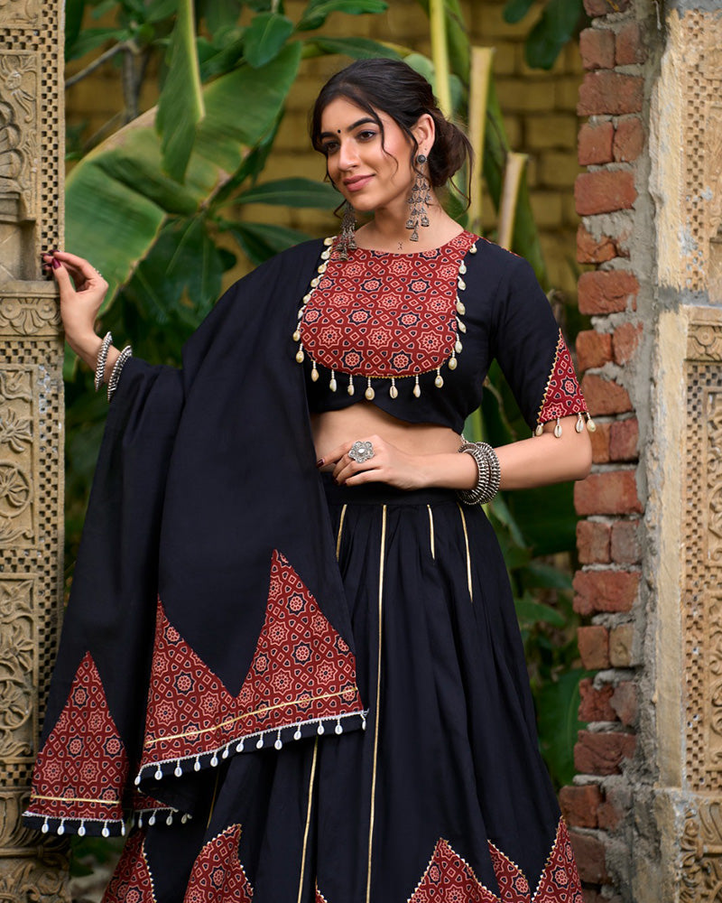 Black cotton printed lehenga choli set with traditional red and black geometric patterns, gota patti and mirror work, paired with a matching dupatta featuring decorative tassels. Ideal for festive occasions or ethnic wear collection, showcased outdoors with ornate stone carvings in the background.