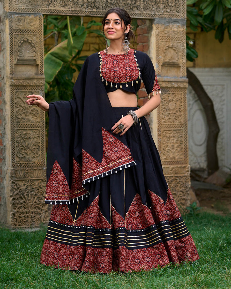 Black cotton printed lehenga choli set with traditional red and black geometric patterns, gota patti and mirror work, paired with a matching dupatta featuring decorative tassels. Ideal for festive occasions or ethnic wear collection, showcased outdoors with ornate stone carvings in the background.