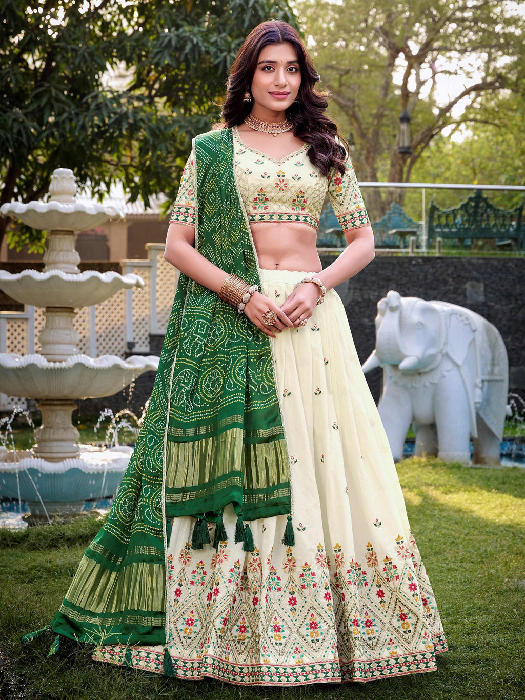 Woman in a green and white traditional outfit standing outdoors with trees and an elephant statue in the background.