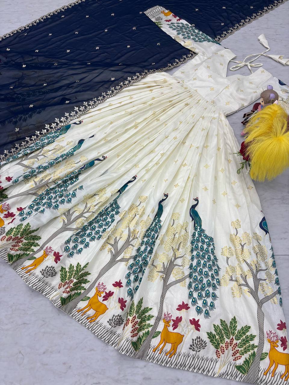 Tarini White Dola Silk Anarkali Gown Set featuring a mesmerizing peacock design with intricate mill print and foil work. Made from premium Dola Silk with a 3-meter flair, it comes with a fully stitched gown and a beautifully embroidered Fox Georgette dupatta adorned with heavy 7mm sequins embroidery. Perfect for festive occasions, weddings, and celebrations, this elegant ensemble blends traditional craftsmanship with contemporary charm