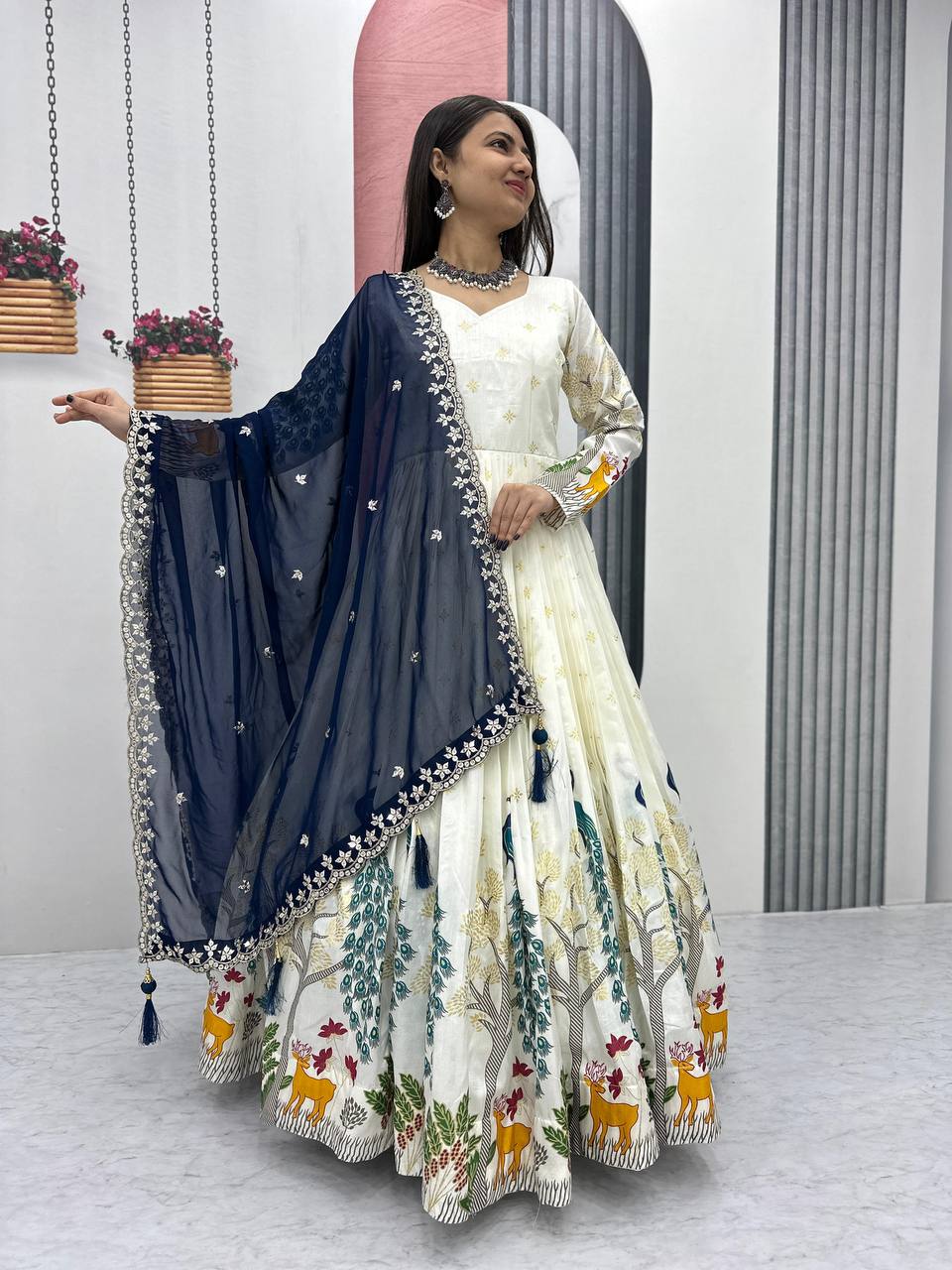 Tarini White Dola Silk Anarkali Gown Set featuring a mesmerizing peacock design with intricate mill print and foil work. Made from premium Dola Silk with a 3-meter flair, it comes with a fully stitched gown and a beautifully embroidered Fox Georgette dupatta adorned with heavy 7mm sequins embroidery. Perfect for festive occasions, weddings, and celebrations, this elegant ensemble blends traditional craftsmanship with contemporary charm